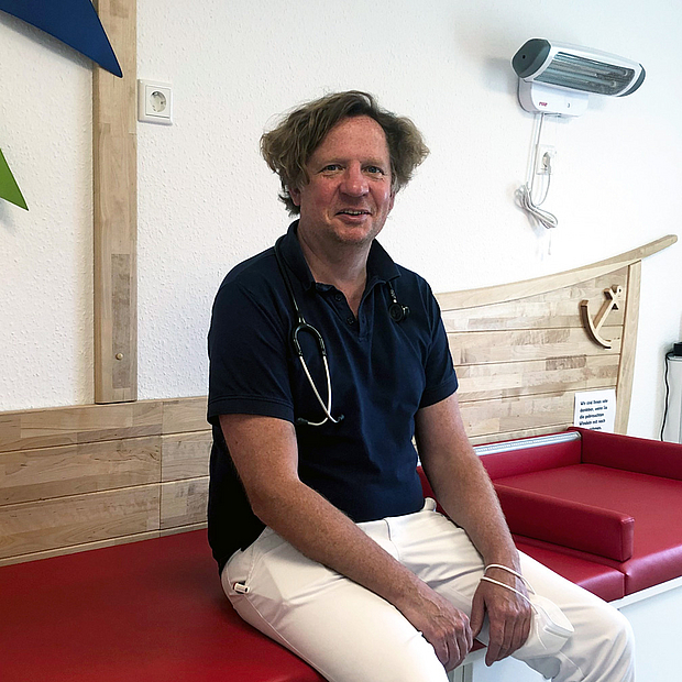 Mann mittleren Alters, Poloshirt, umgehängtes Stethoskop, lächelnd, sitzt auf Behandlungsliege