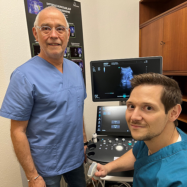 Dr. Jakob Schreiner sitzt vor einem Ultraschallgerät, links steht sein Vater, beide in OP-Kleidung