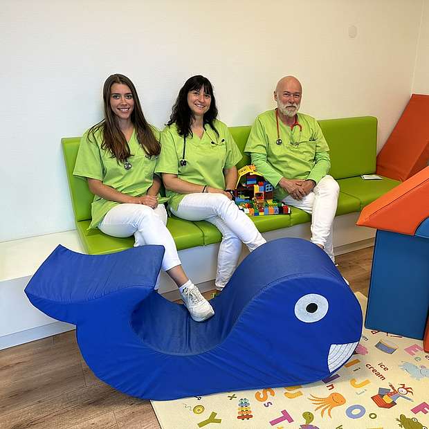 Zwei Frauen und ein Mann sitzen im Wartezimmer einer Kinderarztpraxis.