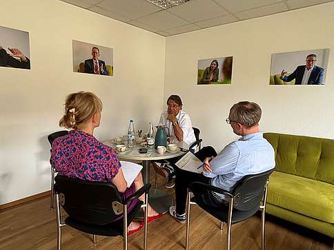 Zu sehen sind zwei Männer und eine Frau, die gemeinsam an einem runden Tisch sitzen. Die Frau in einer der Männer sitzen mit dem Rücken zur Kamera. Der andere Mann unterhält sich mit den beiden. Auf dem Tisch stehen Kaffee und Snacks.